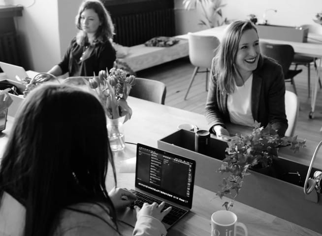 Image of a women sitting at a table smiling, and having a meeting with 2 other women with 1 woman typing on a laptop computer